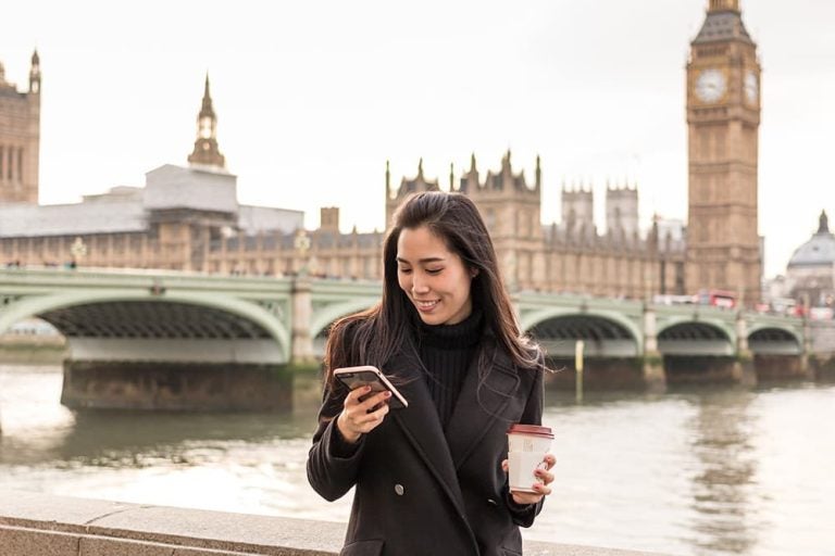 ロンドンのビッグベンを背景に、スマートフォンで通話やメッセージ操作をする女性旅行者