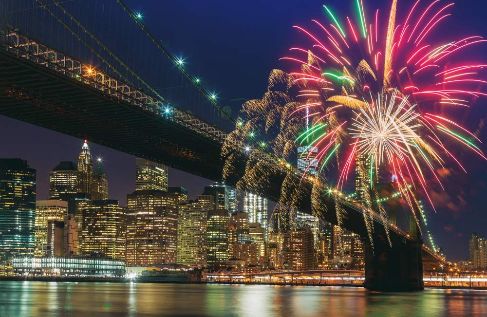 ニューヨークのブルックリン橋を背景に、新年の花火が夜空を彩る壮大な光景。マンハッタンの夜景と水面に映る光が織りなす、カウントダウンクルーズから望む幻想的な瞬間を捉えた一枚。
