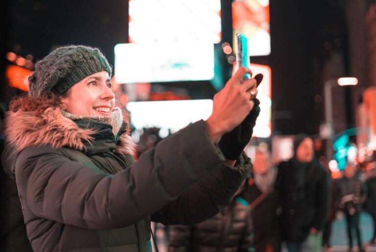 ニューヨーク・タイムズスクエアの夜、ネオンが輝く街並みと混雑する人々を背景に、ニット帽とダウンジャケット着た女性がセルフィ―を撮っている様子