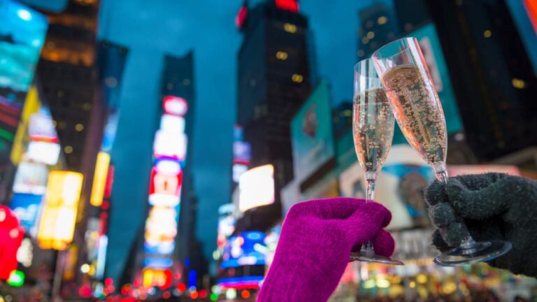ニューヨーク・タイムズスクエアの夜景を背景に、シャンパンを掲げて新年を祝う人々の手元を映した写真。きらめくネオンの中で乾杯する瞬間が、ニューヨークの年越しの華やかさを象徴している。