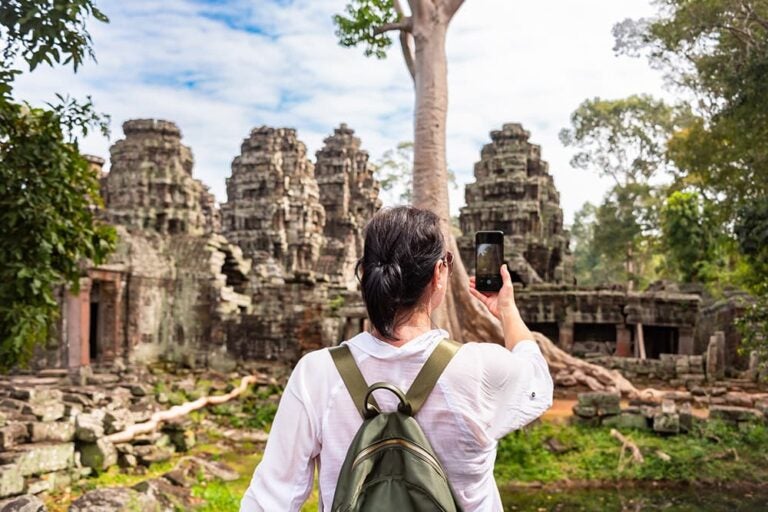 カンボジア・シェムリアップのアンコール遺跡群で、巨木と石造遺跡をバックにスマートフォンで撮影を楽しむ観光客の様子
