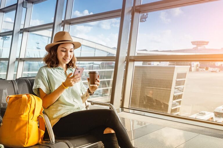 空港の搭乗ロビーでコーヒーを片手にスマートフォンを見ながら、出発を待つ女性旅行者
