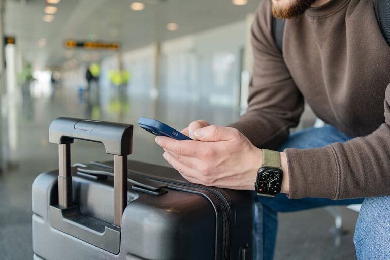 空港でスーツケースの横に座り、出発前にスマートフォンでeSIMの設定を確認する男性