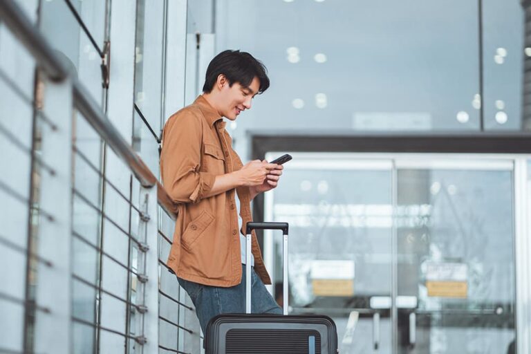 空港でスーツケースの横に立ち、スマートフォンを操作しながら笑顔を見せる男性旅行者