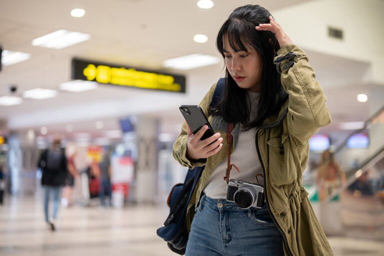 空港でスマートフォンを見ながら、通信や設定がうまくいかず困った表情を浮かべる女性旅行者