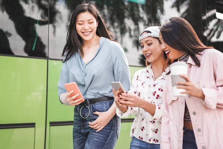 屋外でスマートフォンを見ながら、笑顔で楽しそうに会話する3人の若い女性