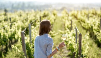 夕暮れのブドウ畑で白ワインを片手に散策する女性の後ろ姿。ボルドー郊外のワイナリーでの穏やかなひとときを表現した一枚