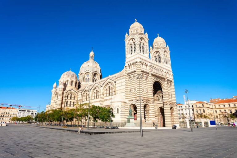 青空の下にそびえるマルセイユ大聖堂の荘厳なビザンチン様式ファサードと開放的な広場の風景