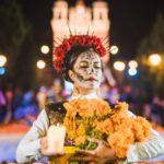マリーゴールドの花束とキャンドルを持ち、カトリーナのメイクをした女性が死者の日の夜祭りを歩く様子