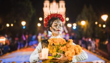 マリーゴールドの花束とキャンドルを持ち、カトリーナのメイクをした女性が死者の日の夜祭りを歩く様子