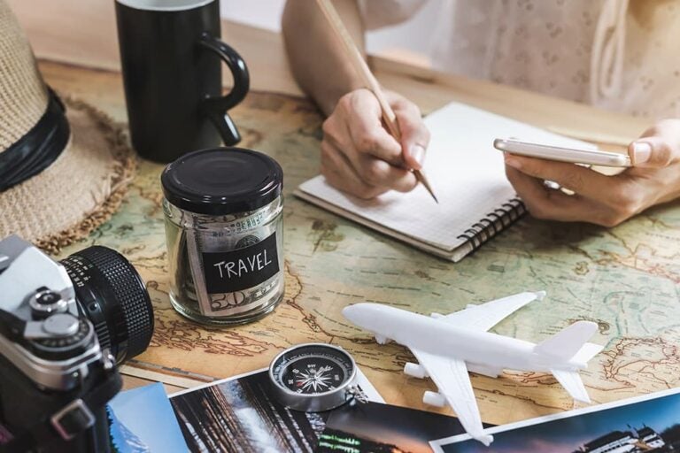 地図の上に旅行貯金の瓶・飛行機の模型・カメラ・コンパスを並べ、スマートフォンを手にノートで旅行計画を立てる様子、Airaloの料金やプランを検討するイメージ