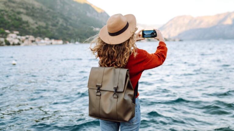 海辺でスマートフォンを構える旅人の後ろ姿と、山並みと水面が広がる風景