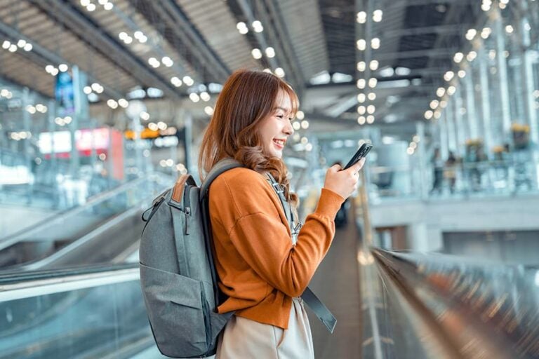 バックパックを背負った女性が空港のターミナルで笑顔でスマートフォンを操作する様子、Airaloがおすすめな旅行者のイメージ
