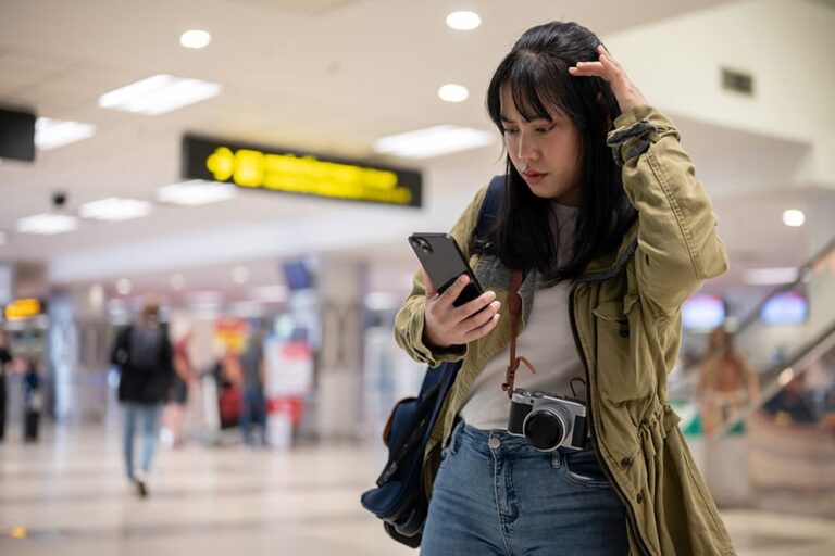 空港のターミナルでカメラを下げた女性が、スマートフォンを見ながら戸惑った表情を浮かべる様子、Airaloを使う前に知っておきたい注意点を示す一枚