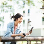 屋外でモバイルバッテリーを使ってスマートフォンを充電しながら楽しそうに操作する女性の様子