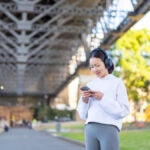 橋の下でスマートフォンを操作しながら音楽を聴くヘッドフォン姿の女性の様子