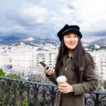 高台から街並みを眺めながらコーヒーとスマートフォンを手に持つ女性