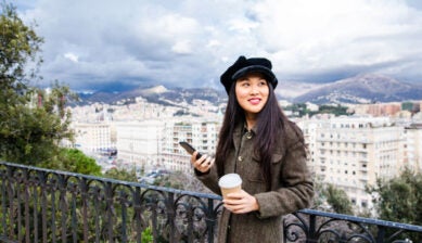 高台から街並みを眺めながらコーヒーとスマートフォンを手に持つ女性