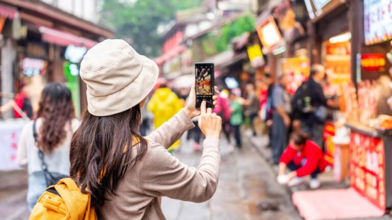 中国の賑やかな街並みで、観光客がスマートフォンを使って屋台や通りの様子を撮影している風景
