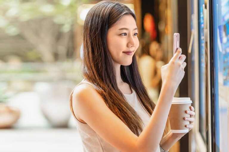 コーヒーを片手に屋外でスマートフォンを操作し、満足そうな表情を見せる女性