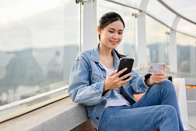 スマートフォンとクレジットカードを手に持ち、屋外でeSIMをオンライン購入・決済する女性