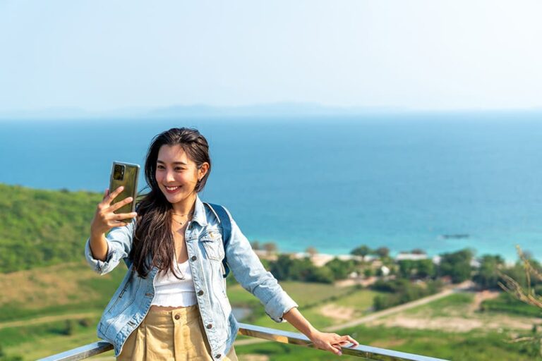 青い海を見渡す高台でスマートフォンを手に、笑顔で旅行を楽しむ女性
