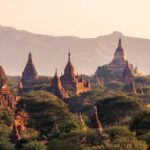 ミャンマーのバガン遺跡群に立ち並ぶパゴダと寺院を、夕暮れの柔らかな光と山並みを背景に望む風景