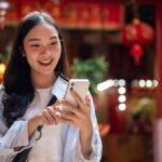 赤い提灯が飾られた寺院の前で、スマートフォンを操作しながら微笑む旅行者の女性