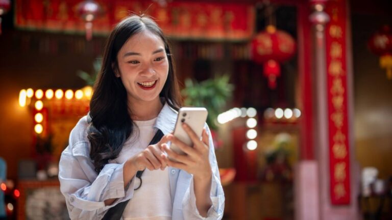 赤い提灯が飾られた寺院の前で、スマートフォンを操作しながら微笑む旅行者の女性