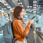 空港のターミナルで、バックパックを背負いながらスマートフォンを操作している旅行者