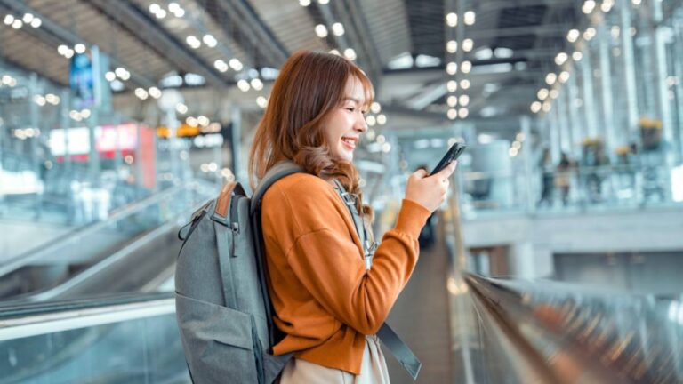 空港のターミナルで、バックパックを背負いながらスマートフォンを操作している旅行者