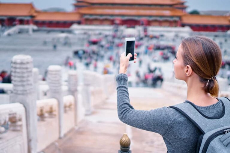  北京・故宮の高台からスマートフォンで広大な宮殿群を撮影する女性旅行者