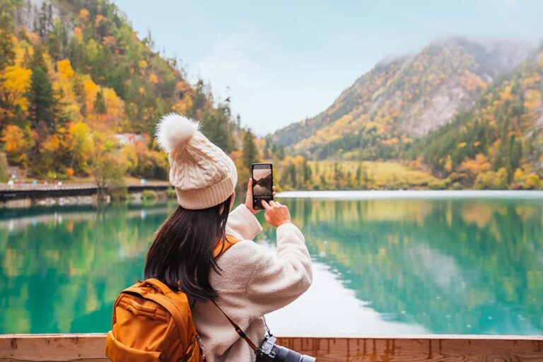 秋の九寨溝でエメラルドグリーンの湖と紅葉の山々をスマートフォンで撮影する旅行者の後ろ姿