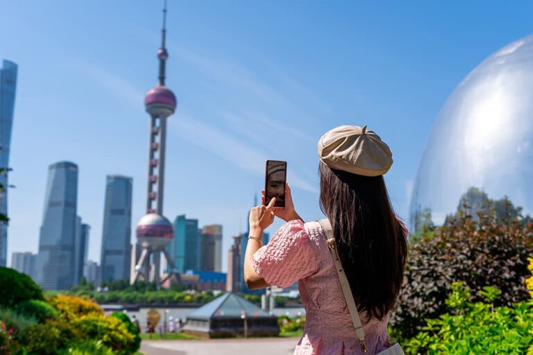 青空の下、上海の東方明珠塔と浦東のビル群をスマートフォンで撮影する旅行者の後ろ姿