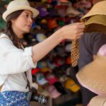 カメラを首から下げた女性旅行者が香港の市場で麦わら帽子を手に取って選んでいる様子