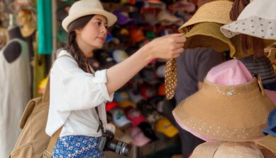 カメラを首から下げた女性旅行者が香港の市場で麦わら帽子を手に取って選んでいる様子