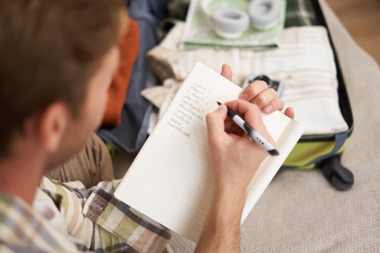 スーツケースの前で旅行の持ち物チェックリストをノートに書く男性のイメージ