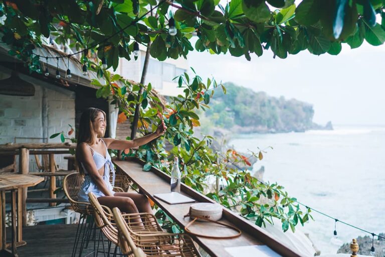 タイの雨季にカフェでスマートフォンを使いながら海を眺める女性旅行者