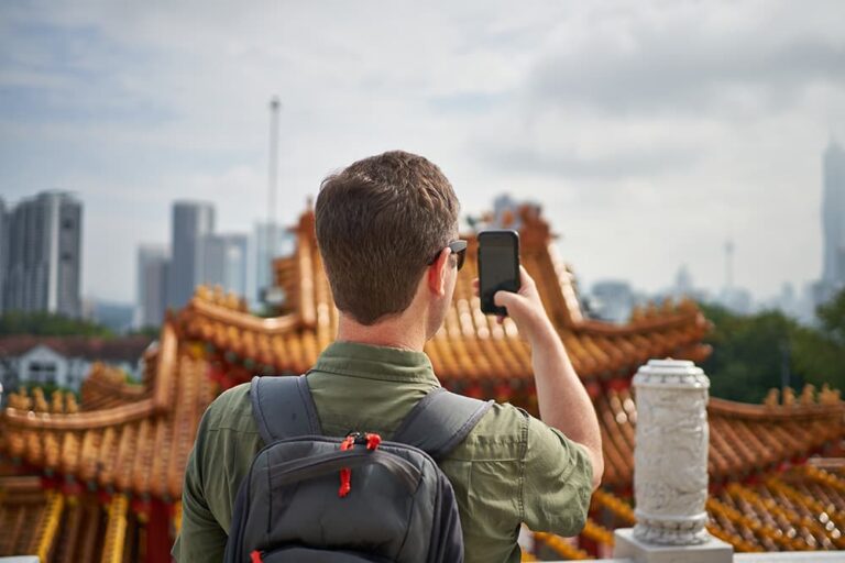 タイの寺院を背景にスマートフォンで撮影する旅行者の後ろ姿。変圧器不要で使える携帯機器の代表例を示す一枚