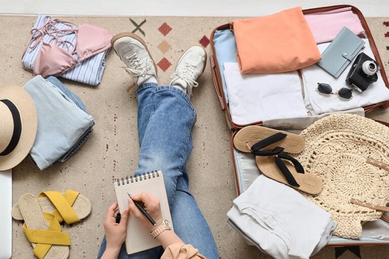 夏服や帽子をスーツケースに詰めながらメモを取る旅行準備の様子