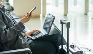 空港の待合エリアでスマートフォンとノートパソコンを使い、テザリングでインターネットに接続するビジネス旅行者