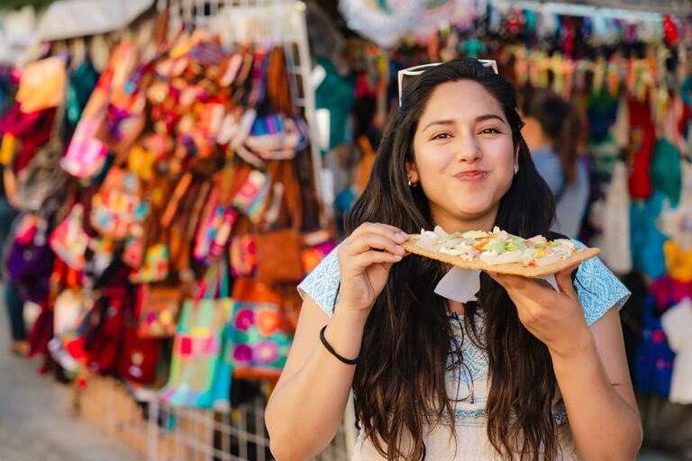 色鮮やかな民芸品が並ぶメキシコの市場でトラユーダを食べる笑顔の女性旅行者