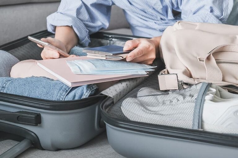 スーツケースの前でスマホとパスポートを手に旅行の準備をする様子。サンディエゴ旅行の出発前の準備を表すイメージ

