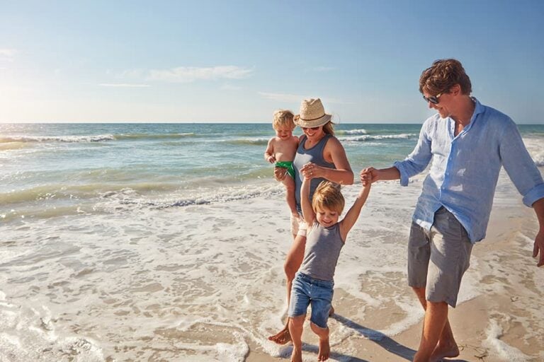 グアムのビーチの波打ち際を手をつないで歩く4人家族。赤ちゃんから幼児まで、子供の成長や年齢に合わせて海辺でのびのびと過ごす様子を捉えた一枚