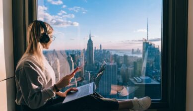 ニューヨークの摩天楼を望む窓辺でノートPCとスマートフォンを使う女性