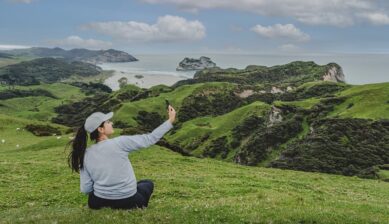 ニュージーランドの丘から海岸の絶景をスマートフォンで撮影する女性