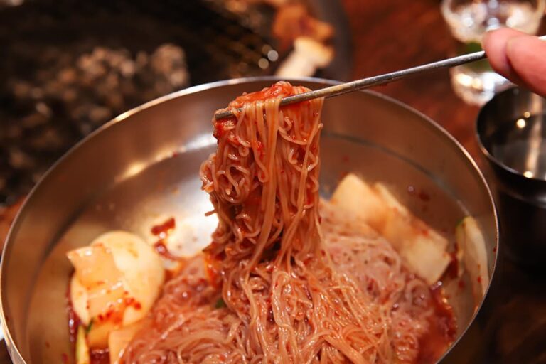 辛いタレを絡めた韓国のビビン冷麺を金属の器から箸で持ち上げている様子