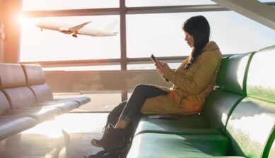 空港の待合席でスマートフォンを操作する旅行者と、窓の外を飛ぶ飛行機