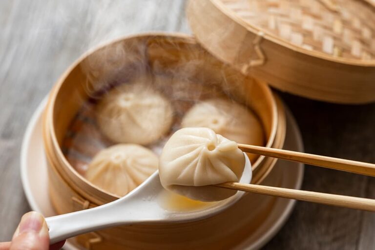 湯気が立ち上るせいろに入った上海名物の小籠包。箸で持ち上げられ、レンゲにたっぷりのスープが溢れ出ている様子