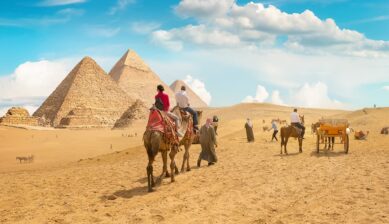 ギザのピラミッドを背景に、砂漠での観光の移動手段としてラクダに乗る人々の様子